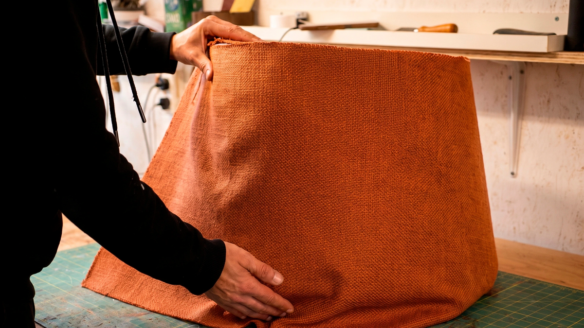 A person in a black hoodie holds up a large piece of textured orange-brown woven fabric in a workshop setting.